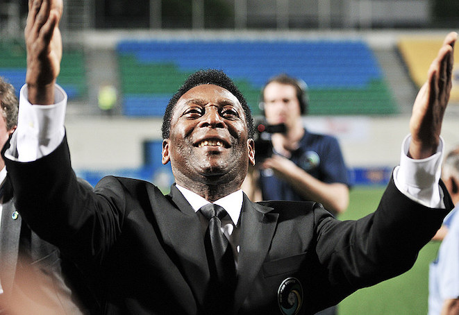 Pele, a Black man, standing with his arms up as he looks up to the stands in a soccer stadium
